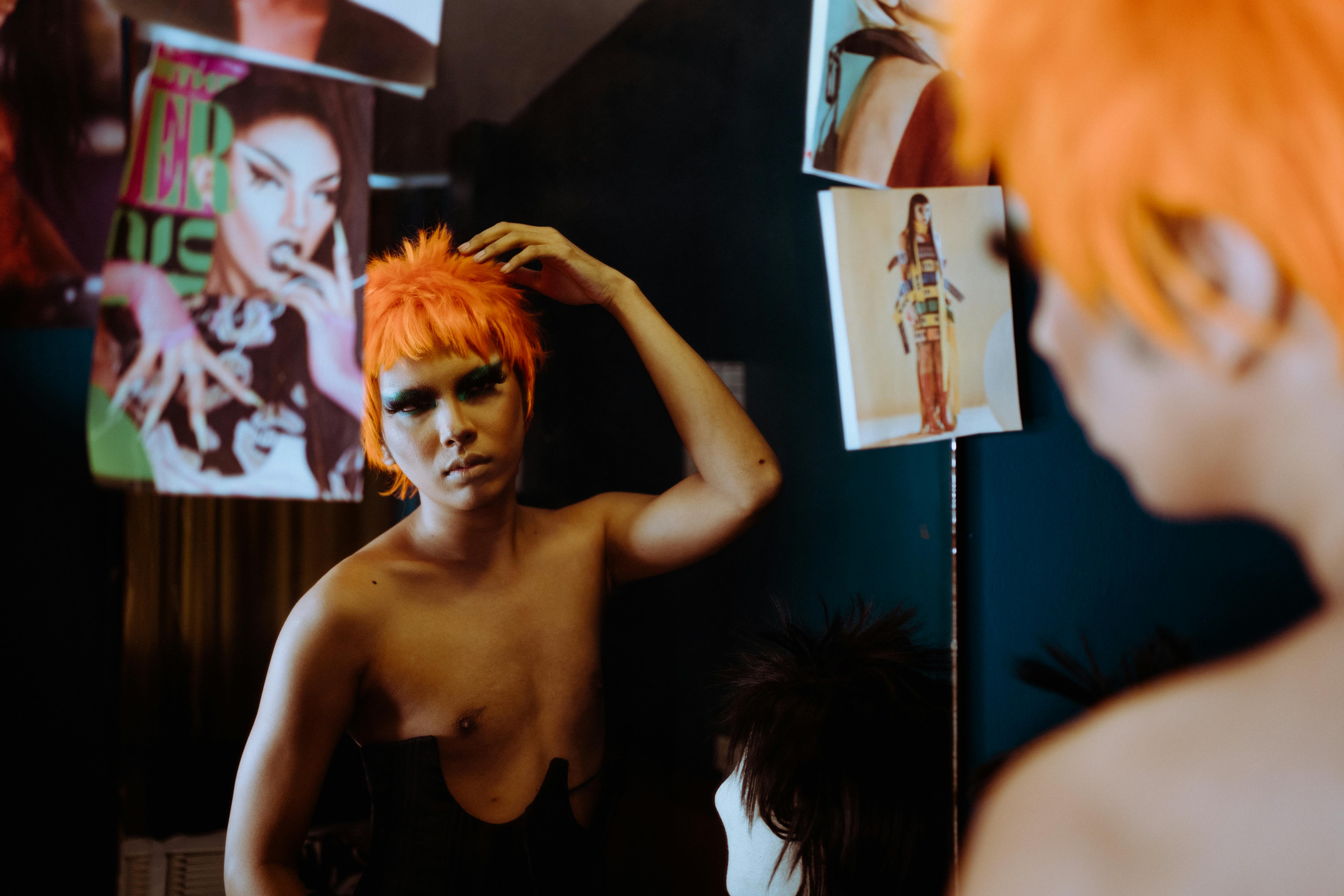 Free Drag performer adjusts wig in a mirror, surrounded by photos, highlighting art and transformation. Stock Photo