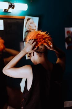 A person styles a bright orange wig in front of a mirror, embodying bold fashion creativity.