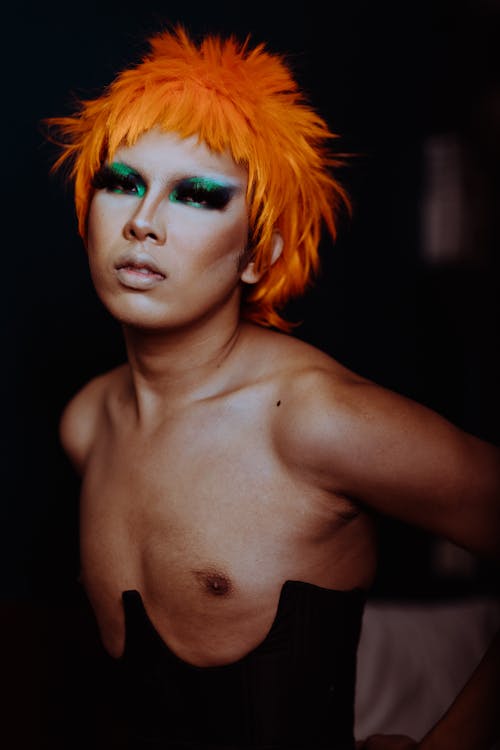 Young ethnic androgynous man with cool shiny makeup and bare chest in corset looking at camera