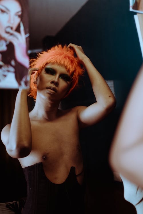 Provocative young ethnic androgynous guy with makeup in black corset and orange wig looking in mirror in dressing room