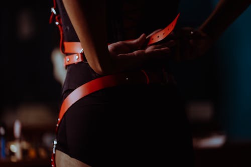 Back view of crop unrecognizable female in black corset and panties fastening leather harness in dressing room