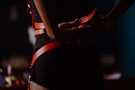 Back view of crop unrecognizable female in black corset and panties fastening leather harness in dressing room