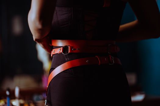 Back view of crop anonymous female in black panties corset and leather belt standing in dressing room and looking in mirror