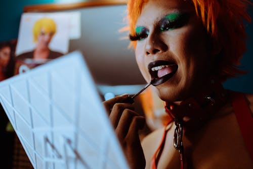 Free Crop eccentric ethnic androgynous male with artistic makeup and orange wig looking in mirror and applying lipstick Stock Photo