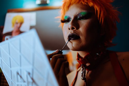 Free Low angle of crop ethnic eccentric transgender guy with bright makeup in wig and leather collar applying dark lipstick while preparing for performance in nightclub Stock Photo