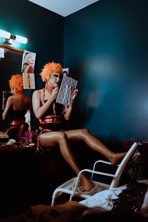 Full body of young informal androgynous male artist in eccentric stage clothes and wig sitting on mirror table in dressing room and applying makeup before performance
