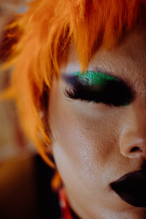Closeup of crop eccentric ethnic androgynous person with bright artistic makeup in orange wig standing with closed eyes