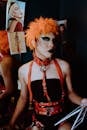 Confident young ethnic transsexual guy with bright makeup in orange wig and BDSM harness and collar sitting in dressing room and looking at camera before show in nightclub