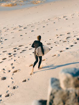 Surfer in wetsuit carrying surfboard on sandy Bronte Beach in Australia, leaving footprints in the sand.