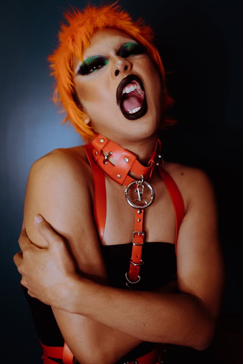 Free Naughty young ethnic androgynous guy with bright makeup in BDSM collar and wig embracing self and looking at camera with opened mouth Stock Photo