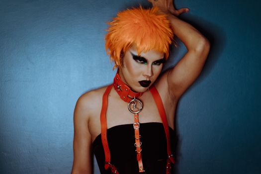 Serious young ethnic transgender male model with makeup in BDSM collar and wig touching blue wall and looking away in studio