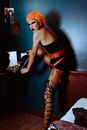 Side view of provocative young ethnic gender fluid male with bright makeup in eccentric BDSM outfit and orange wig tying shoelaces in bedroom and looking at camera