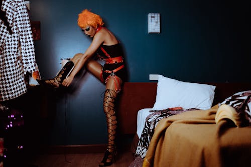 Side view of eccentric young ethnic transvestite with makeup in BDSM costume and wig wearing high heeled shoes in bedroom
