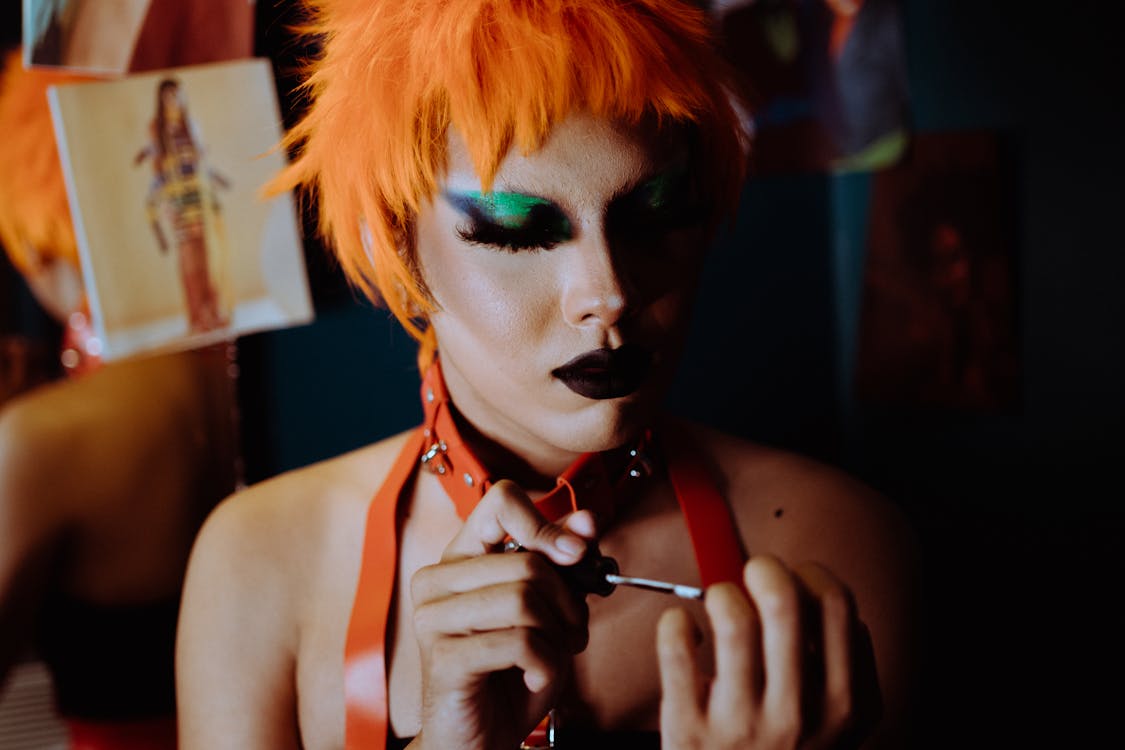 Crop eccentric female with orange dyed hair and bright makeup wearing leather harness and painting nail