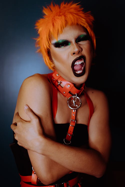 Expressive drag queen in orange wig and with dark makeup wearing leather harness embracing self and opening mouth eccentrically while looking at camera