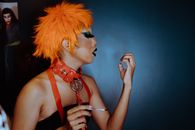 Side view transgender male with orange dyed hair and dark makeup blowing on painted nails while standing against dark blue wall in studio