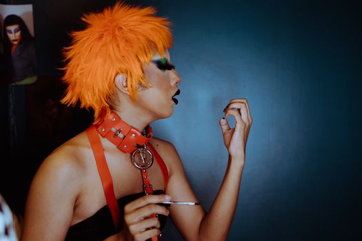 Side view transgender male with orange dyed hair and dark makeup blowing on painted nails while standing against dark blue wall in studio