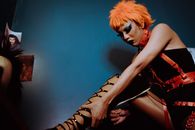 Side view slim transsexual guy in BDSM clothes and with makeup fastening zipper on thigh high shoes