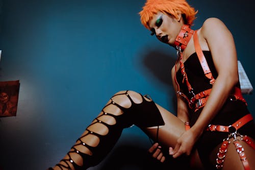 Free From below transgender guy model with dark makeup and dyed hair raising leg and fastening high lace up shoe in dark room Stock Photo