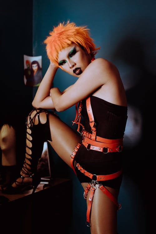 Free Side view of confident ethnic transsexual guy in provocative BDSM accessories and bodysuit leaning on hand and looking at camera in dressing room Stock Photo