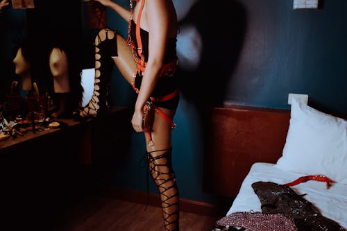Free Side view of crop unrecognizable transsexual male in eccentric BDSM harness and bodysuit standing with leg on mirror table in bedroom Stock Photo