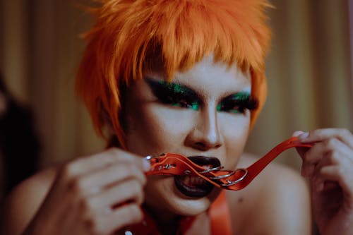 Crop provocative ethnic female with aggressive makeup in orange wig biting leather collar and looking at camera