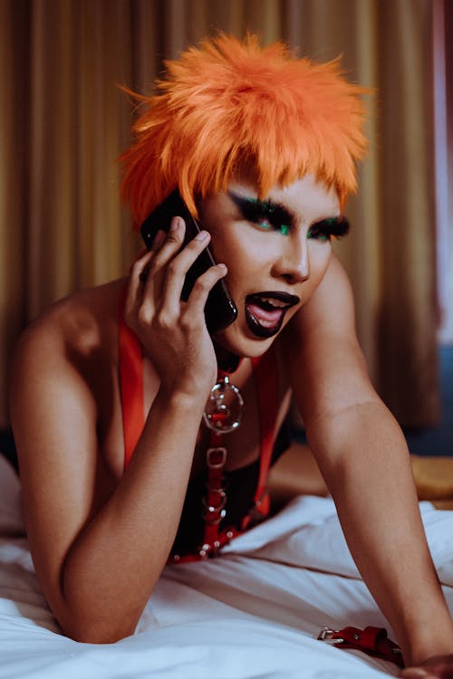 Free Provocative young ethnic androgynous male with bright makeup in wig lying on bed and looking at camera while having phone conversation Stock Photo