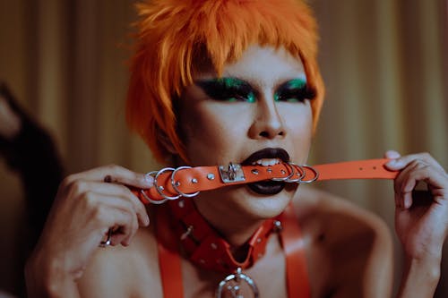 Free Crop seductive young ethnic woman with bright makeup in orange wig and BDSM collar lying on bed and biting red leather strap Stock Photo