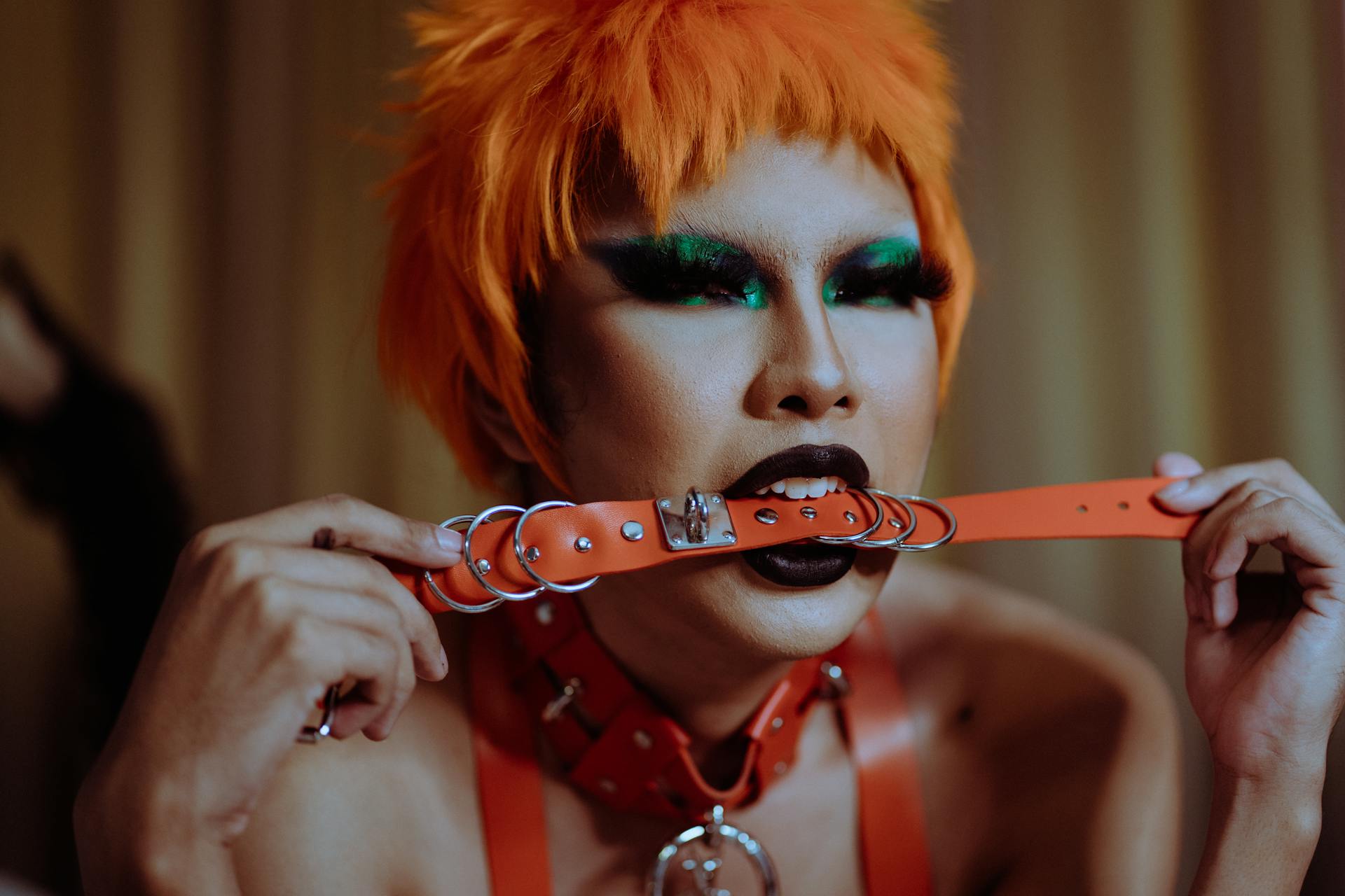 Crop seductive young ethnic woman with bright makeup in orange wig and BDSM collar lying on bed and biting red leather strap