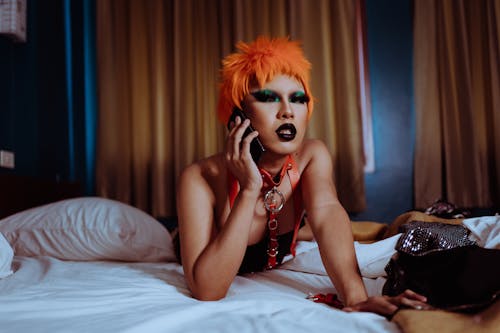 Free Seductive young ethnic androgynous male with bright makeup in orange wig and BDSM collar lying on soft bed on stomach and talking on mobile phone Stock Photo