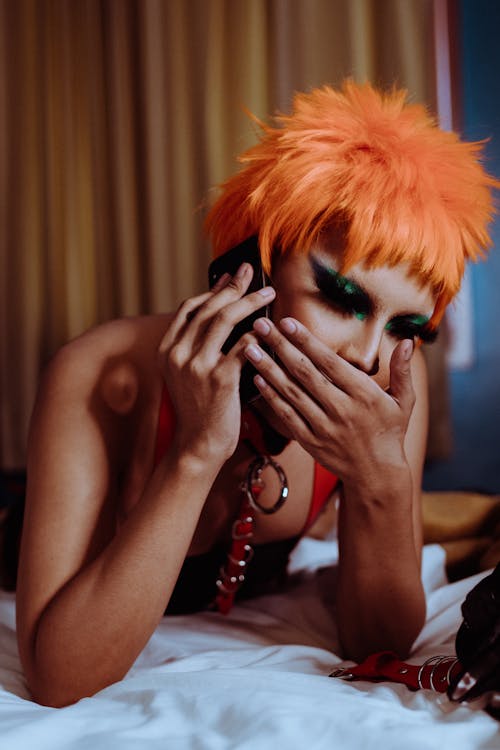 Free Eccentric young ethnic androgynous person with bright makeup in wig covering mouth with hand while lying on bed and talking on mobile phone Stock Photo