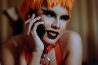 Crop young informal ethnic lady with bright makeup and short orange hair smiling while lying on stomach and talking on smartphone