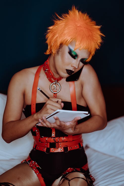 Free Serious young ethnic androgynous guy with aggressive makeup in provocative BDSM costume and orange wig sitting on bed and taking notes in diary during phone conversation Stock Photo