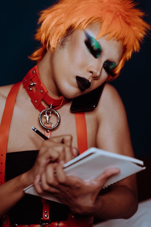 Crop focused ethnic actress with bright makeup in wig talking on cellphone while taking notes in notepad