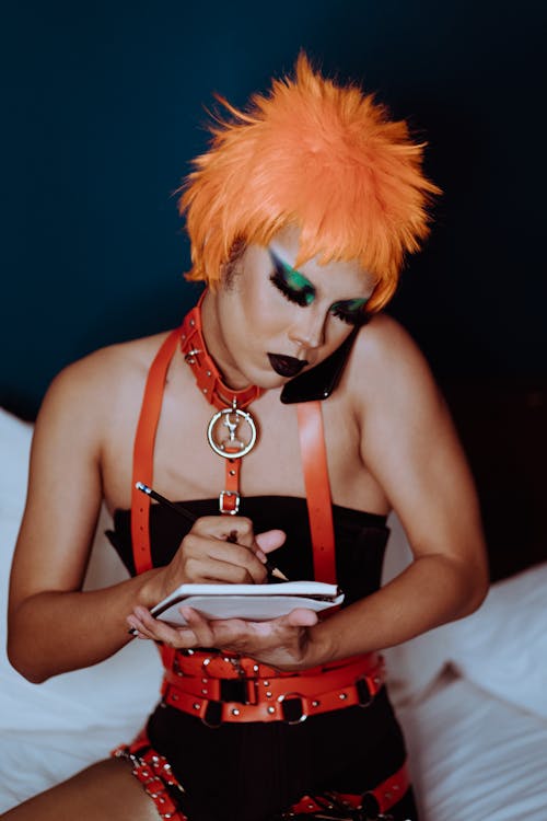Young female artist with bright makeup in wig and bondage talking on mobile phone while taking notes in notebook on bed