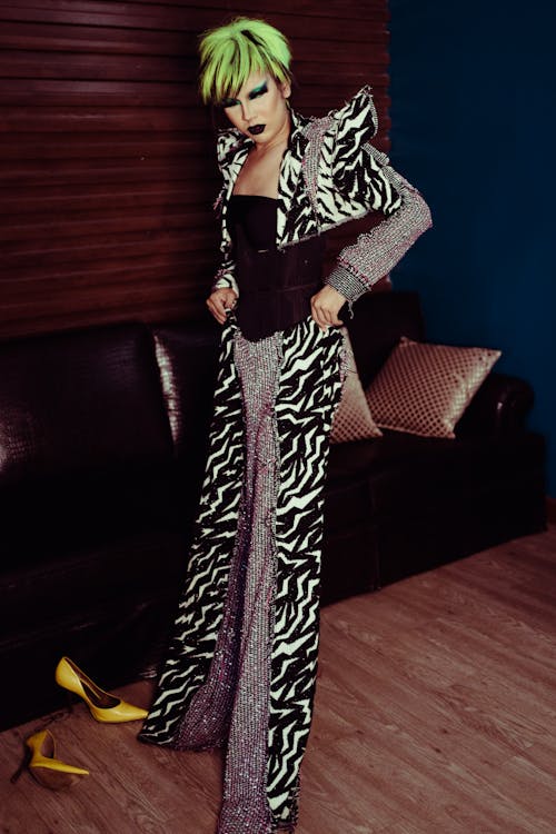 Young ethnic female theater artist with makeup in wig putting on ornamental trousers on parquet in dressing room