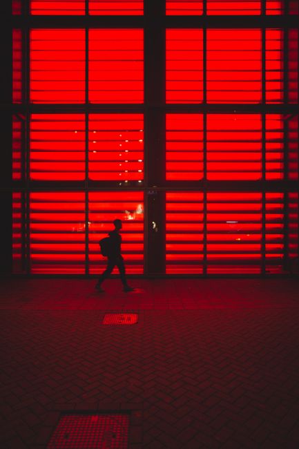 A lone figure walks past a vividly red illuminated wall, creating a striking silhouette.