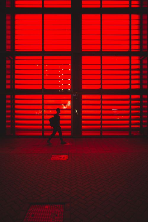 Free A lone figure walks past a vividly red illuminated wall, creating a striking silhouette. Stock Photo