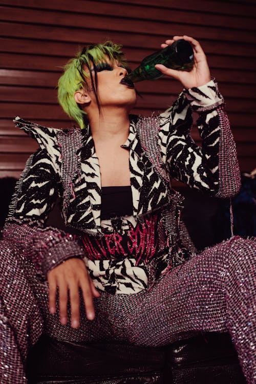 Transsexual man with colorful hair and bright makeup in stylish outfits sitting on sofa and drinking bottle of beverage in apartment