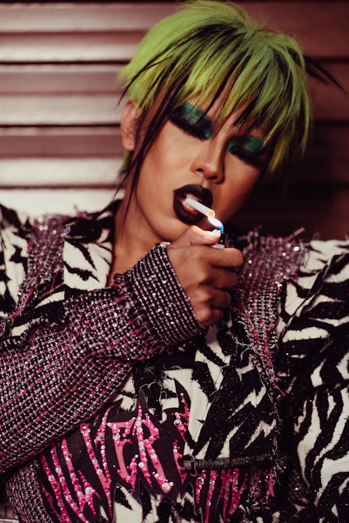 Confident transgender model with colorful makeup and bright hairstyle in festive stylish clothes lighting cigarette