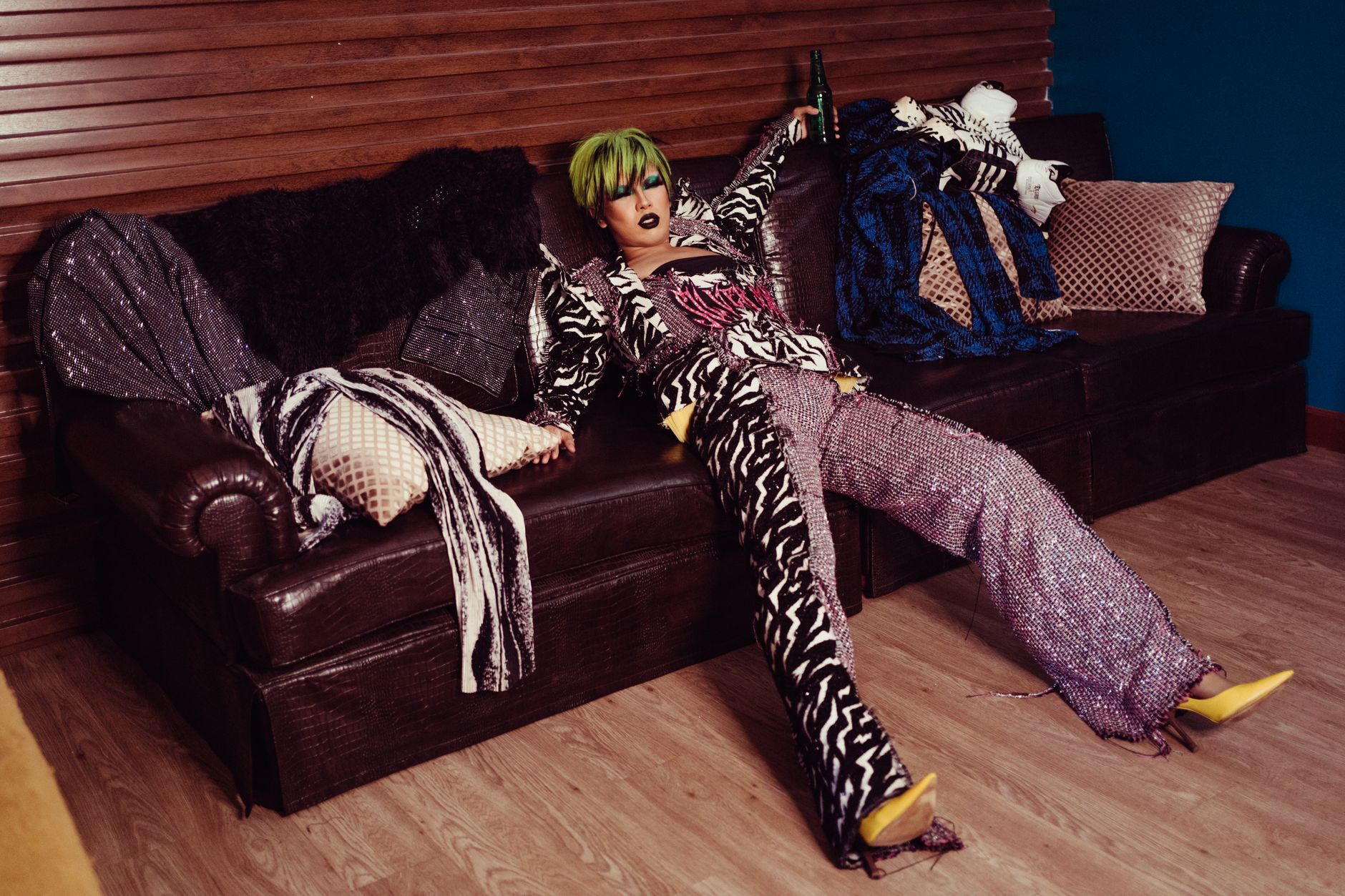 Full body of extravagant androgynous model with creative makeup and hairstyle in festive stylish clothes resting with bottle of beverage on leather sofa in apartment
