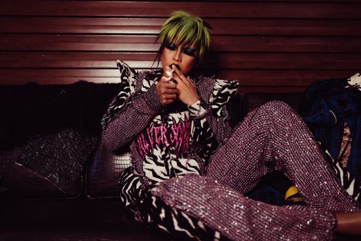 Stylish male transsexual with bright makeup and trendy outfit with glitter relaxing on couch while smoking cigarette