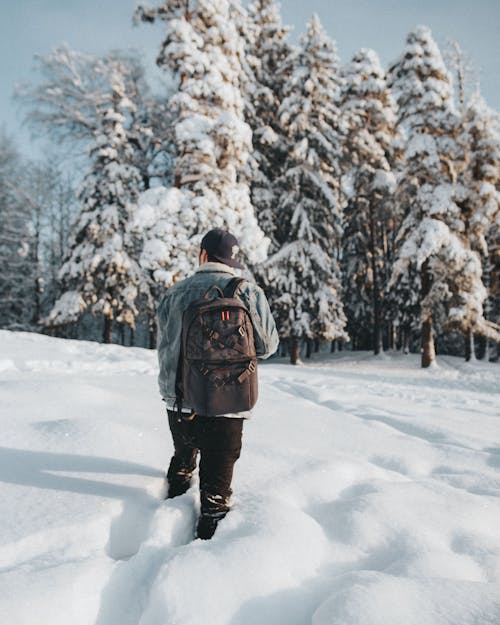 Man in Denim Jacket and Black Pants Walking on Snow-Covered Ground
