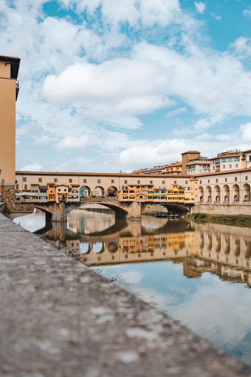 Free A scenic view of the Ponte Vecchio bridge reflecting on the River Arno in Florence under a vibrant blue sky. Stock Photo