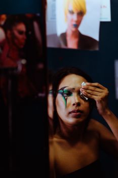 Young transgender with long hair removing makeup on face in front of mirror with pictures