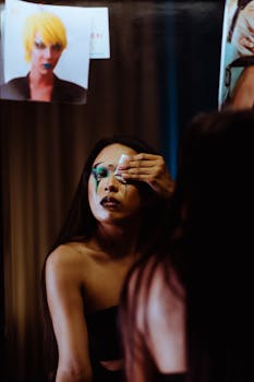 Young crossdresser with long dark hair tissuing eye while removing makeup standing in front of mirror