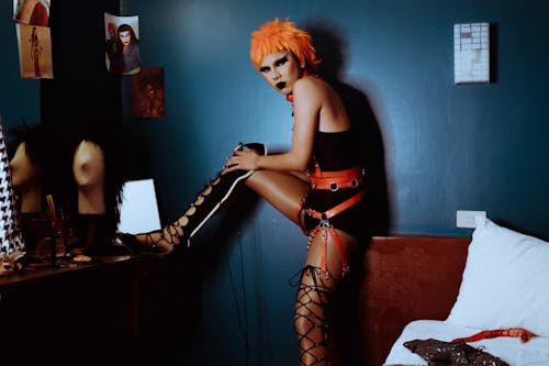 Side view unemotional extravagant female with orange dyed hair and dark makeup wearing bodysuit and thigh high shoes putting leg on vanity table and looking at camera