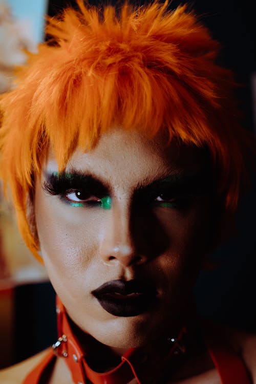 Transsexual young female with provocative makeup wearing orange wig and leather harness looking at camera confidently