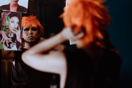 Back view crop provocative female with bright makeup in black dress putting on orange wig and looking in mirror