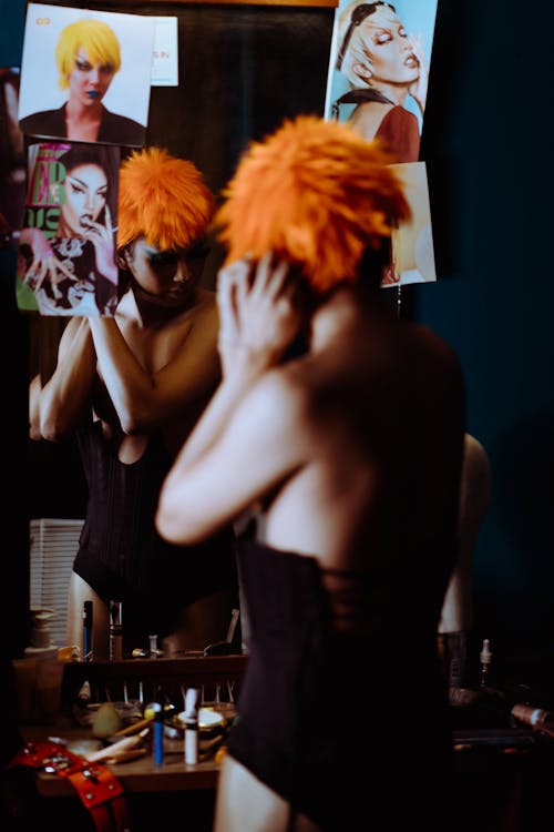 Back view bizarre female in black bodysuit putting on bright orange wig against mirror in dressing room